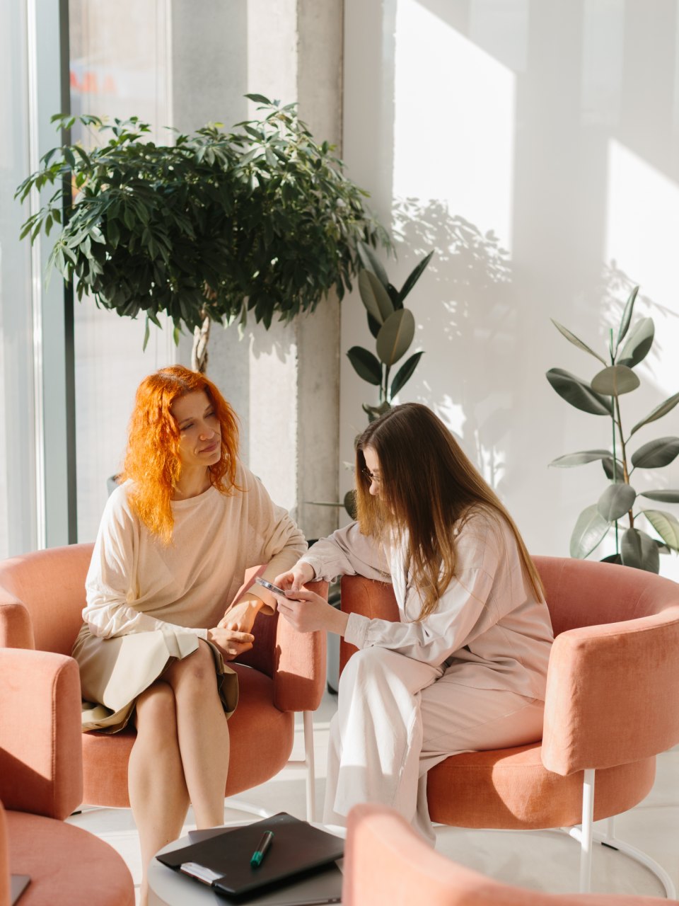 Two women talking face to face in peach-coloured armchairs, beside a large houseplant