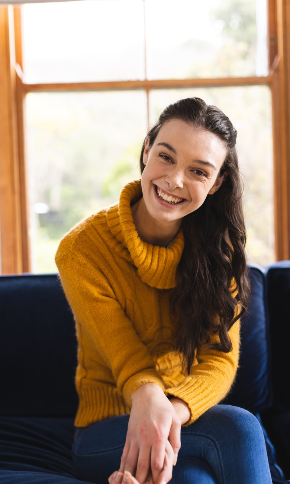 A smiling woman in a yellow jumper sitting in front of a window in a bright room